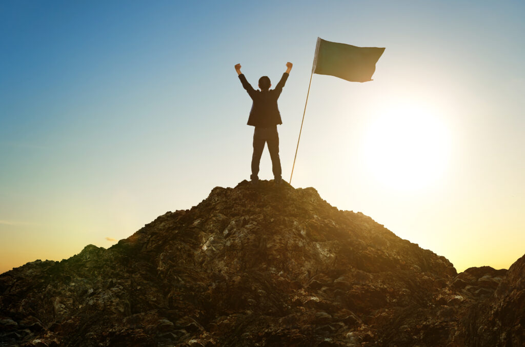 graphicstock-business-success-leadership-achievement-and-people-concept-silhouette-of-businessman-with-flag-on-mountain-top-over-sky-and-sun-light-background_SOllvyhPxjg Silhouette of a leader with a flag on a mountain top at sunrise, symbolising positivity and thoughtful leadership built over years